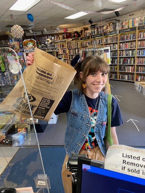 A customer holding a printed Bag Ad Agency campaign bag at the counter of an independent comic book shop